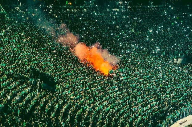 Luftaufnahme einer großen Menge von Menschen in einem Stadion mit einem zentralen Feuer, das die Umgebung erhellt.