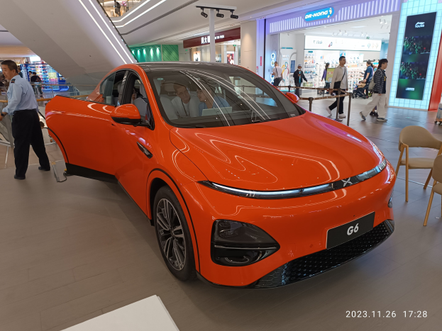 Orangefarbenes Elektroauto in einer Ausstellungshalle mit einer Gruppe von Menschen drumherum, umgeben von Stühlen, einem Wagen, Geschäften, Namensschildern und Lichtern im Hintergrund.