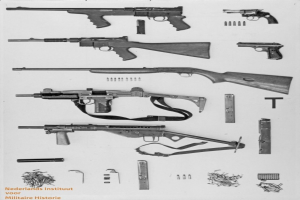 Schwarz-weiß-Foto von verschiedenen Feuerwaffen, einschließlich einer Maschinengewehr, mit verstreuter Munition und Gegenständen und Text unten.