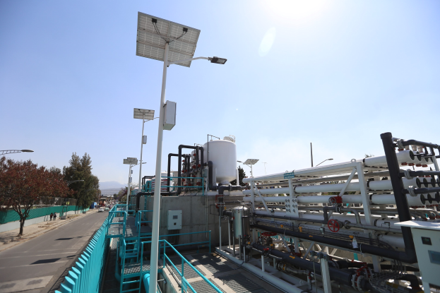 Großes industrielles Gebäude mit ausgedehnten Rohrleitungen und Solarpanelen, umgeben von Sicherheitsgeländern, Strommasten, Straßenlaternen, Schildern, Bäumen und vorbeifahrenden Fahrzeugen, vor einem Hügel- und einem klaren blauen Himmel, Teil einer kalifornischen Wasseraufbereitungsanlage.