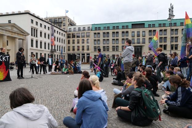 Eine Gruppe von Menschen, die auf dem Boden vor einer Menge sitzen, die Fahnen und Transparente schwenkt, bei einer Demonstration gegen Homosexuelle in Berlin, mit einer Statue, Gebäuden und einer Person, die in ein Mikrofon spricht.