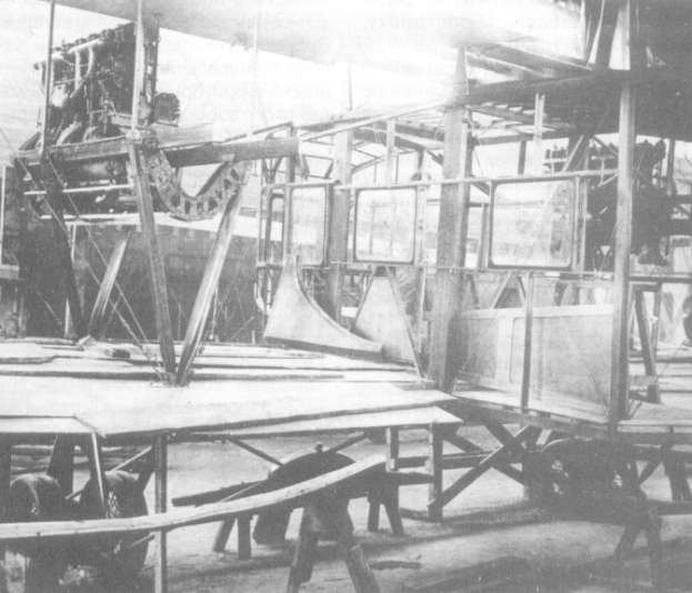 Ein Schwarz-Weiß-Foto eines Flugzeugs in der Fertigung in einer Fabrik, umgeben von Metallstangen und anderen Gegenständen.