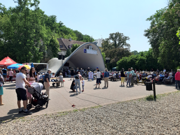 Menschenmenge vor einem Gebäude auf einem Festival Opening, einige sitzen auf Bänken, andere stehen, einschließlich eines Kinderwagens im Vordergrund; Hintergrund zeigt Bäume, Gebäude und einen klaren blauen Himmel, mit Zelten links und einem Müllcontainer rechts.