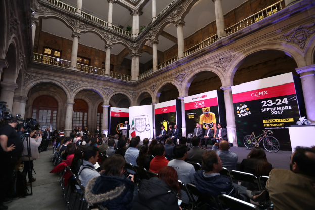 Eine Gruppe von Menschen, die an einem Tisch mit einem Podium und einem Mikrofon vor einer Menge, Kameras und Transparenten sitzen, was auf eine Pressekonferenz für das CDMX 2017-Event hindeutet.