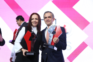 Zwei Personen auf einer Bühne, die strahlend Trophäen halten, mit einem Bildschirm im Hintergrund