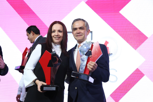 Zwei Personen auf einer Bühne, die strahlend Trophäen halten, mit einem Bildschirm im Hintergrund