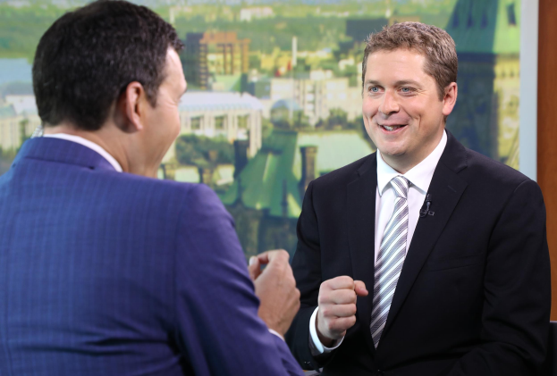 Ein Mann in einem blauen Anzug, weißem Hemd und gemustertem Schlips sitzt in einem Stuhl und hat die Hände verschränkt, während er mit einem anderen Mann in einem professionellen Setting mit einem gerahmten Objekt an der Wand hinter ihnen spricht.