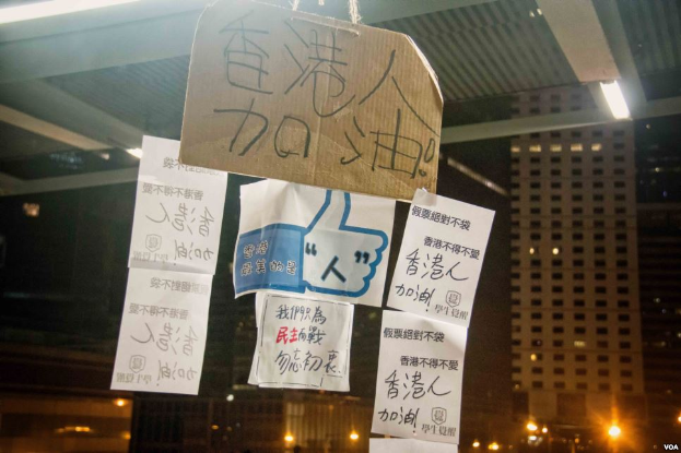 Eine Gruppe von Menschen steht nachts vor einem Gebäude, das von Stadtlichtern beleuchtet wird, mit mehreren Protestschildern an der Decke.