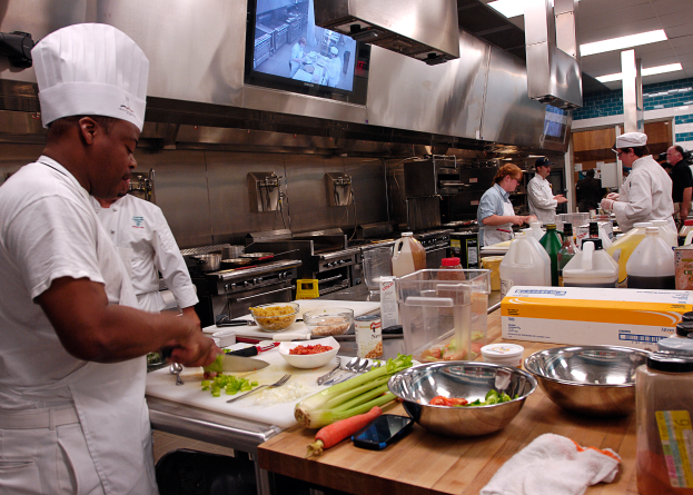 Eine Gruppe von Köchen in einer Küche, mit einem im Vordergrund in einer weißen Schürze und Mütze, der ein Messer hält und von verschiedenen Kochutensilien, Gemüse und anderen Gegenständen auf dem Tisch umgeben ist, mit einem Fernseher und Deckenleuchten im Hintergrund.