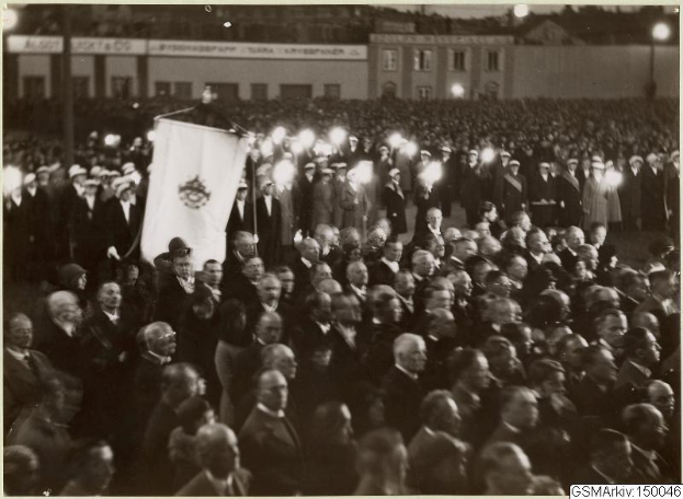 Schwarzes und weißes Foto einer großen Menschenmenge vor einem Gebäude, mit einer Person, die eine Fahne hält, sichtbaren Masten und Lichtern im Hintergrund und einem Text unten, der "Gustav Klimts Rede an der Berliner Mauer" lautet.