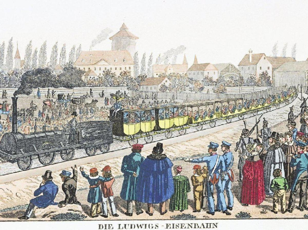 Eine Gruppe von Menschen, die um einen Zug auf Schienen an der ersten Eisenbahnstation in Deutschland stehen, mit Gebäuden und Bäumen im Hintergrund.