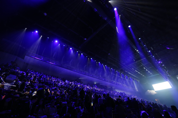 Eine große Menschenmenge sitzt einer hell erleuchteten Bühne mit deckenmontierten Scheinwerfern gegenüber, mit einem Bildschirm auf der rechten Seite, was auf eine Konzerthalle hinweist.