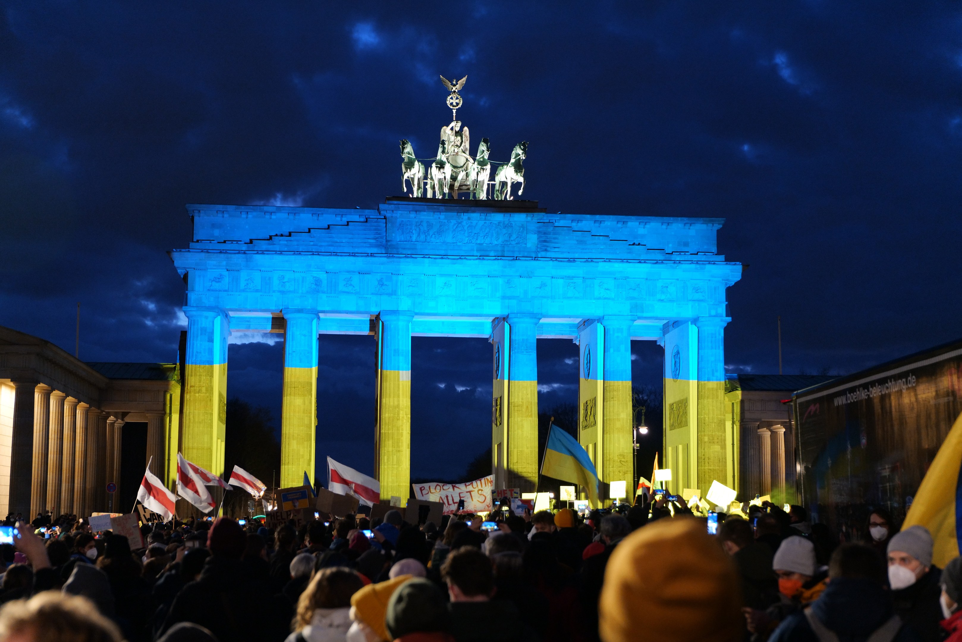 Eine Menschenmenge steht vor dem Brandenburger Tor in Berlin und hält Fahnen und Schilder in den Händen, mit einer Fahne auf der rechten Seite und bewölktem Himmel darüber.