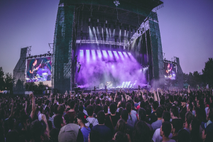 Eine große Menschenmenge versammelt sich auf einem Musikfestival mit einer Bühne, auf der Musiker Instrumente spielen, beleuchtet von Lichtern und umgeben von Bildschirmen, vor einer Kulisse aus einem Gebäude, Bäumen und einem klaren blauen Himmel.