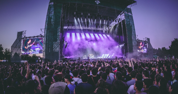 Eine große Menschenmenge versammelt sich auf einem Musikfestival mit einer Bühne, auf der Musiker Instrumente spielen, beleuchtet von Lichtern und umgeben von Bildschirmen, vor einer Kulisse aus einem Gebäude, Bäumen und einem klaren blauen Himmel.