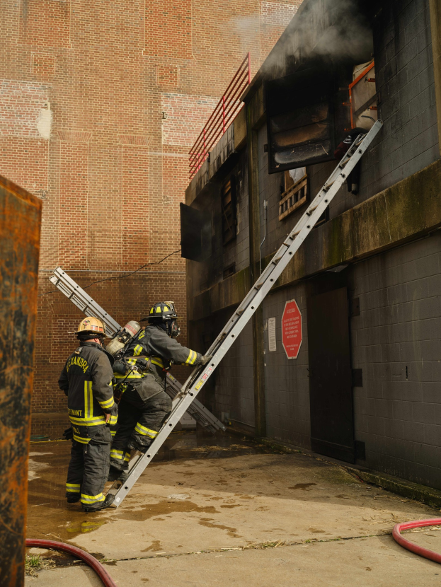 Feuerwehrleute mit Helmen klettern eine Leiter zu einem rauchgefüllten Gebäude hoch, mit einem Metallobjekt links und Rohren auf dem Boden.