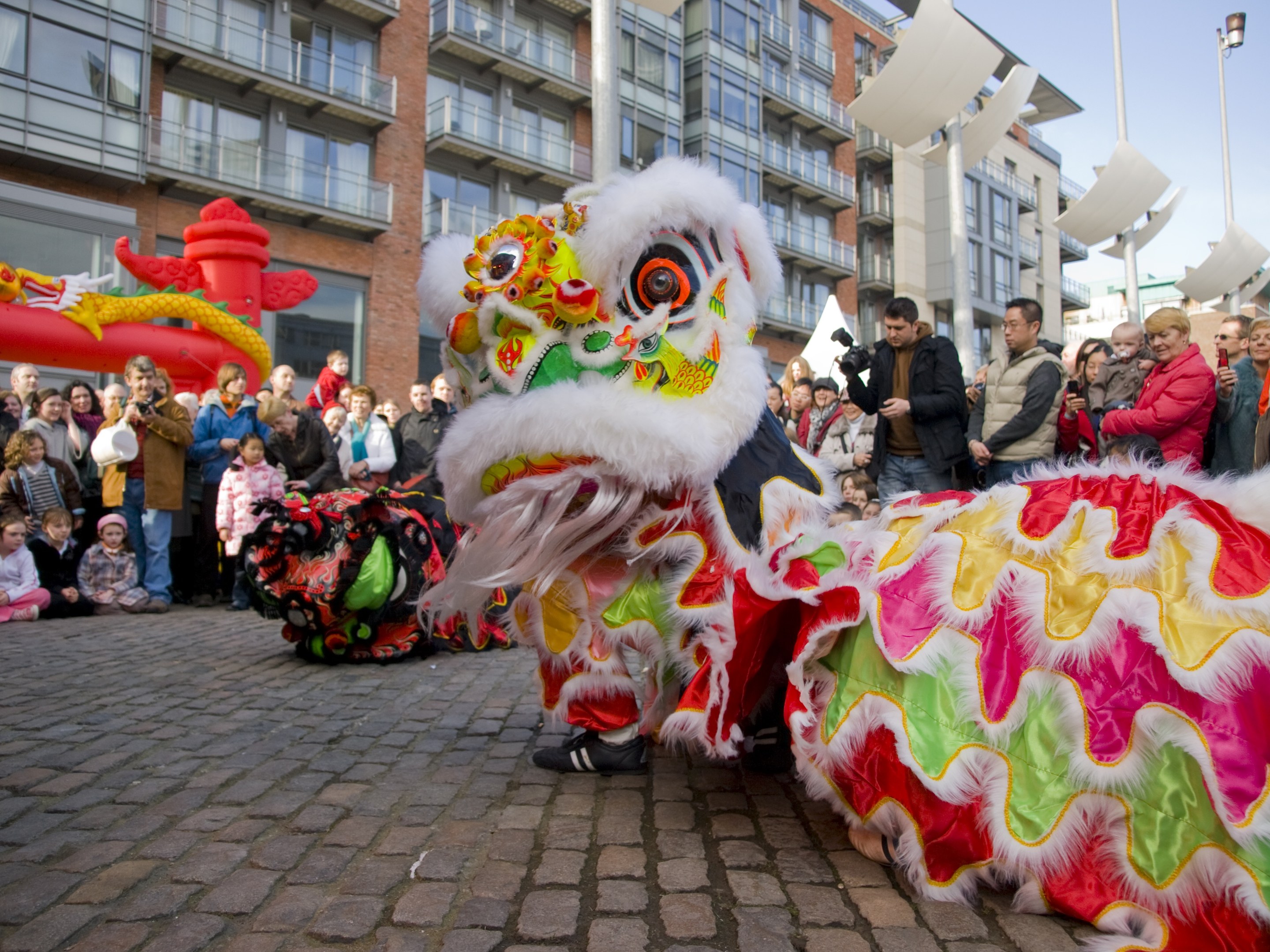 Ein lebendiges chinesisches Neujahrsfest in Amsterdam mit einem Löwen tanzen im Vordergrund und einer Menge Menschen drumherum, einige halten Kameras, vor Gebäuden, Laternenmasten und einem klaren blauen Himmel.