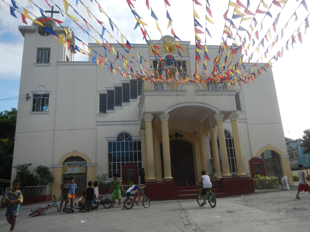 Gruppe von Menschen, die vor einer weißen Kirche mit architektonischen Details wie Fenstern, Säulen, Bögen, Türen und einem Kreuzsymbol Fahrrad fahren, mit Pflanzen, einem Zaun, einer Fahne, Bäumen und einem bewölkten Himmel im Hintergrund und bunten Fahnen, die an einem Seil oben befestigt sind.