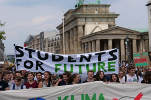 Gruppe von Studenten marschiert in Berlin mit einem bunten 'Students for Future'-Plakat an Gebäuden, Bäumen und Himmel vorbei.