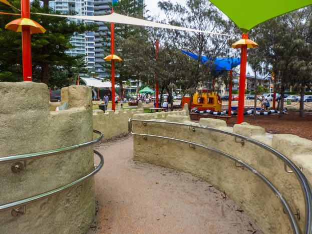 Ein Spielplatz mit einem grünen und gelben Schirm, umgeben von Geländern, Pfählen, Zelten, Bäumen, Fahrzeugen und Menschen, mit Gebäuden und einem klaren blauen Himmel im Hintergrund.