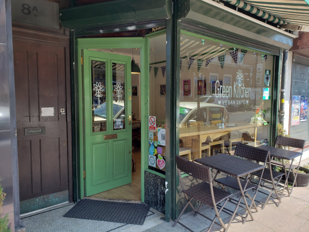 Ein veganes Restaurant namens Green Kitchen Vegan Cafe mit Außenbereich, Topfpflanzen, einer Fußmatte, einer Glastür mit Postern und Innenräumen mit Tischen, Stühlen und Beleuchtung.