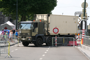 Ein Lkw am Straßenrand mit einer Gruppe von Menschen in der Nähe, umgeben von städtischer Infrastruktur und Grünflächen unter einem klaren blauen Himmel.