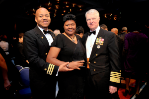 Zwei Männer und eine Frau auf einem roten Teppich, die Frau hält eine Trophäe, mit anderen im Hintergrund und einer beleuchteten Decke.