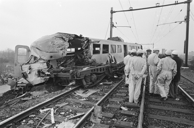 Schwarzes und weißes Bild eines Zugunglücks auf den Schienen mit Einsatzkräften in Helmen und einem Hintergrund aus Strommasten, Bäumen, Gebäuden und einem klaren Himmel.