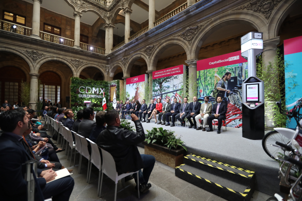 Eine Gruppe von Menschen sitzt auf Stühlen und schaut auf einen Mann auf der Bühne, der ein Mikrofon hält, rechts stehen Fahrräder und im Hintergrund sind Plakate, Pflanzen, Säulen, Geländer, Lampen, Fenster und eine Wand zu sehen.