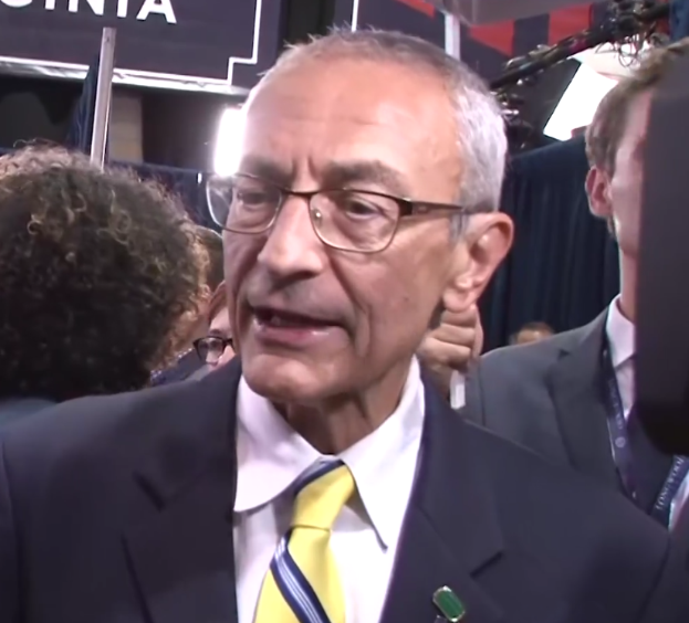 Ein Mann im Anzug und Krawatte spricht zu Reportern, trägt eine Brille und steht hinter einem Mikrofon mit Ständer, umgeben von einer Gruppe von Menschen, mit Lampen und einer Tafel im Hintergrund.