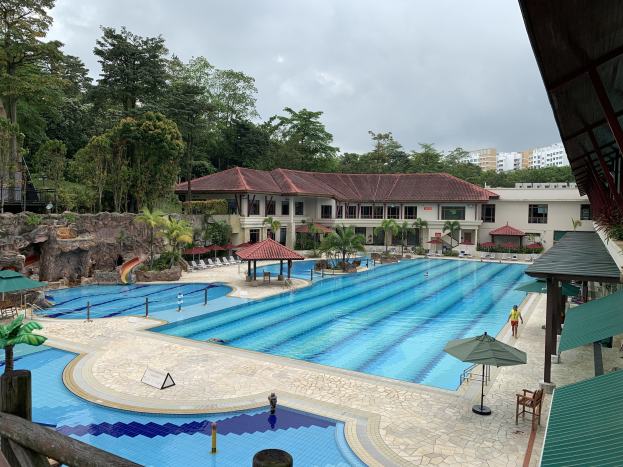 Großes Schwimmbad im Ritz-Carlton, Kota Kinabalu, mit Menschen, Sonnenschirmen, Liegestühlen, Tischen, Pflanzen, Bäumen, Felsen, Gebäuden und einem Schuppen sowie Himmel im Hintergrund.