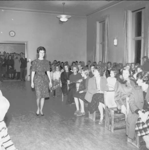 Eine Frau in einem schwarzen und weißen Kleid läuft über einen Laufsteg während einer Modenschau für die Frauenwahlrecht, mit einer Menge, die sitzt und zusieht, einer Uhr und Lampen an der Wand und einer beleuchteten Decke.