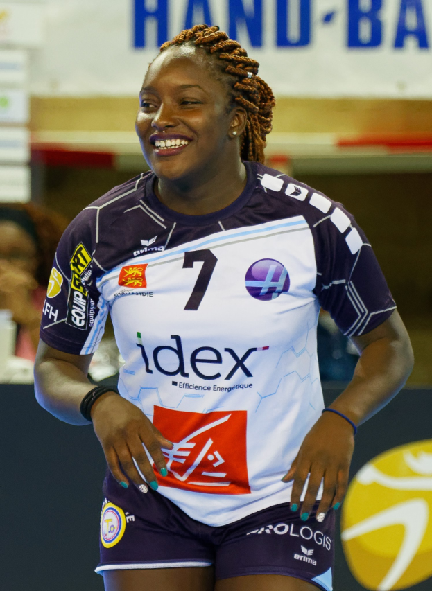 Professionelle Handballspielerin in lila-weißer Uniform, die selbstbewusst auf einem Feld lächelt, mit "FIVB Handball-Weltmeisterschaft 2019"-Schild im Hintergrund.