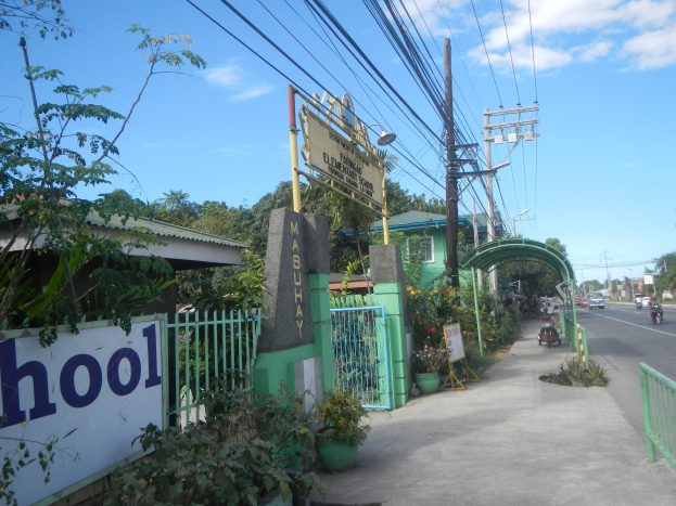 Einfahrt zu einer Schule mit einem "Schule"-Schild am Straßenrand, Fahrzeuge, ein Tor, ein Zaun, eine Namensplakette, Pflanzen, Bäume, Strommasten mit Drähten und einen bewölkten Himmel.