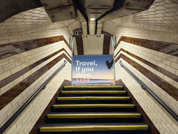Eine Treppe in einer U-Bahn-Station mit Geländern auf beiden Seiten, die zu einem Schild führt, auf dem "Reise, wenn du wagst" steht, mit Stationwänden im Hintergrund.