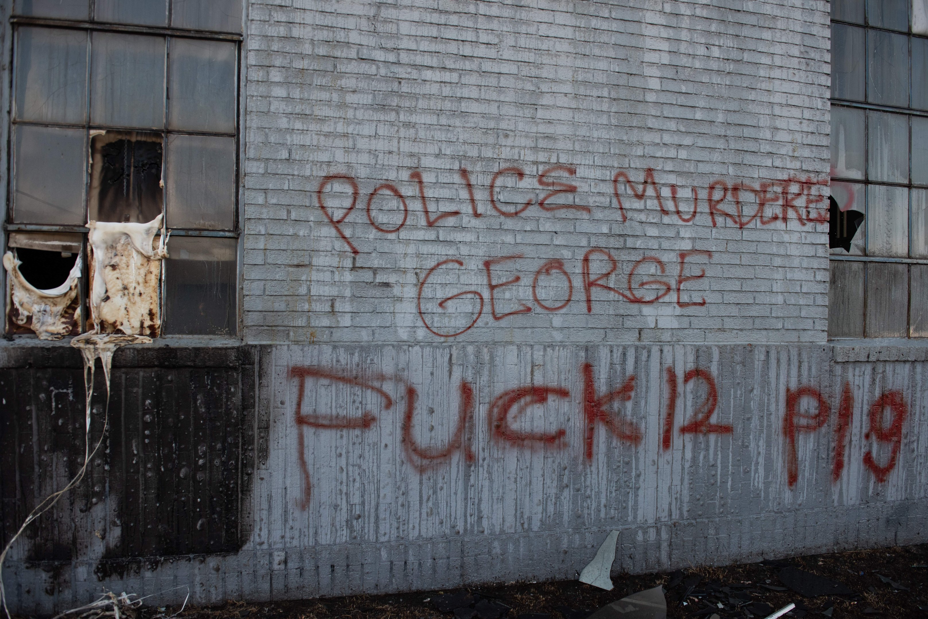 Graffiti auf einer Gebäudewand mit der Aufschrift 'Polizei ermordet George Fuck 12 P19' mit Fenstern und verstreuten Gegenständen davor.