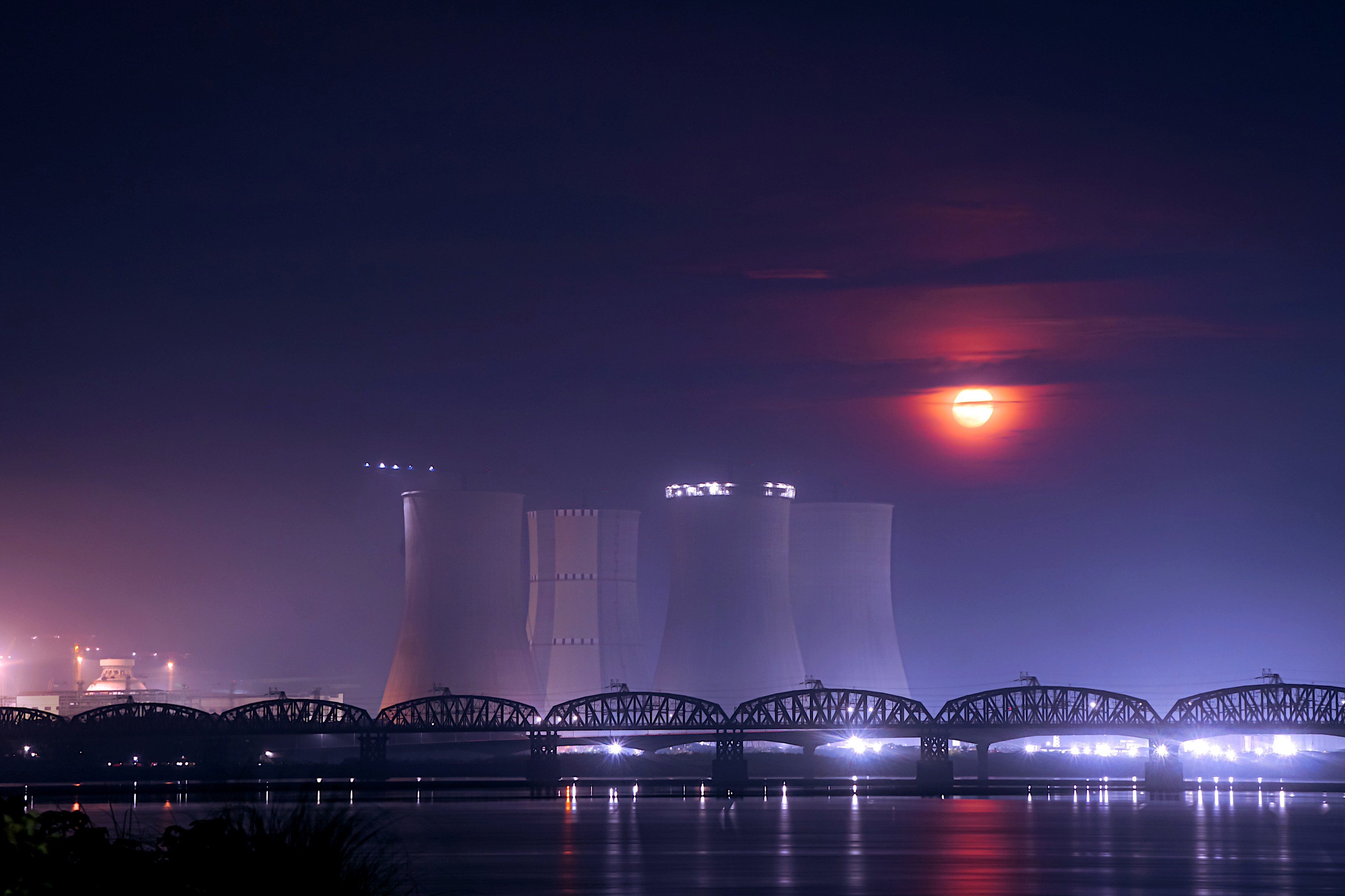 Atomkraftwerk bei Nacht mit Brücke über Wasser, beleuchtet von Gebäudelichtern und Mond, umgeben von Bäumen.