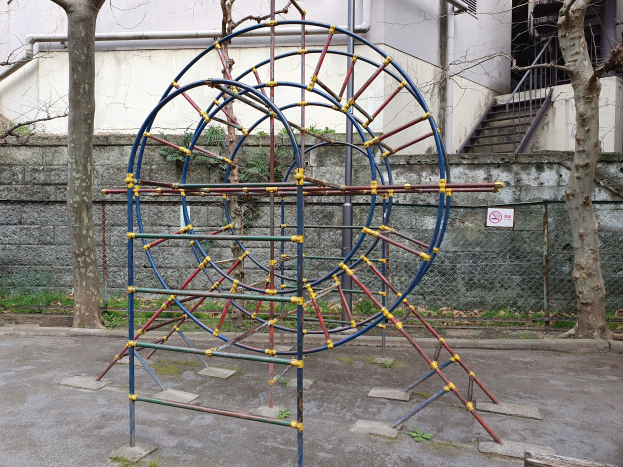 Spielplatz mit einem großen Metallklettergerüst, umgeben von Bäumen und einer Maschendrahtzaunwand, mit Treppen mit Geländern, die zu einem Gebäude mit Fenstern und Rohren führen, einem Schild an einem Pfahl und grasbewachsenem Boden.