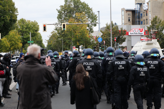 Eine Gruppe von Menschen geht eine Straße entlang, wobei Polizeibeamte anwesend sind, einige tragen Helme und tragen Taschen oder Kameras, mit Fahrzeugen, Verkehrsampeln, Schildern, Bäumen, Gebäuden und einem klaren blauen Himmel im Hintergrund.