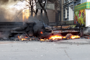 Ein Lkw brennt am Straßenrand, mit Rauch, der aus ihm aufsteigt, während im Hintergrund Bäume, Gebäude und ein klarer Himmel zu sehen sind.
