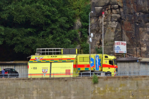 Rettungswagen auf der Seite einer Straße neben einer Klippe geparkt, mit verschiedenen Gegenständen wie einer Wand, einem Geländer, Pfosten, Lampen, Schildern mit Text im Vordergrund und Bäumen und einem Auto im Hintergrund.