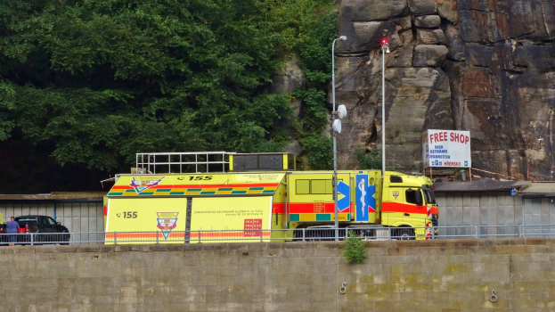 Rettungswagen auf der Seite einer Straße neben einer Klippe geparkt, mit verschiedenen Gegenständen wie einer Wand, einem Geländer, Pfosten, Lampen, Schildern mit Text im Vordergrund und Bäumen und einem Auto im Hintergrund.