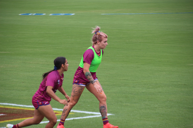 Zwei Frauen spielen Fußball auf einem Feld, eine trägt ein grünes T-Shirt und Shorts, die andere ein pinkes Kleid. Beide tragen Sportkleidung und haben Tätowierungen an Händen und Füßen.