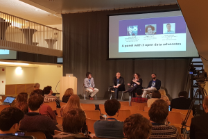 Eine Gruppe von Menschen sitzt auf Stühlen und schaut auf einen Projektionsschirm, auf dem ein Laptop steht. Rechts im Bild steht eine Kamera auf einem Stativ, im Hintergrund sind Geländer, Lampen und eine Wand zu sehen. Die Menschen diskutieren über offene Datenaktivisten.