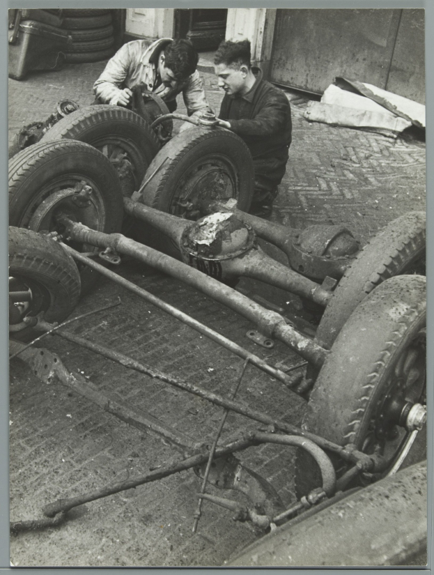 Zwei Männer in einer schwarzen und weißen Garage reparieren ein Auto und halten Werkzeuge, umgeben von Reifen und anderen verstreuten Gegenständen.