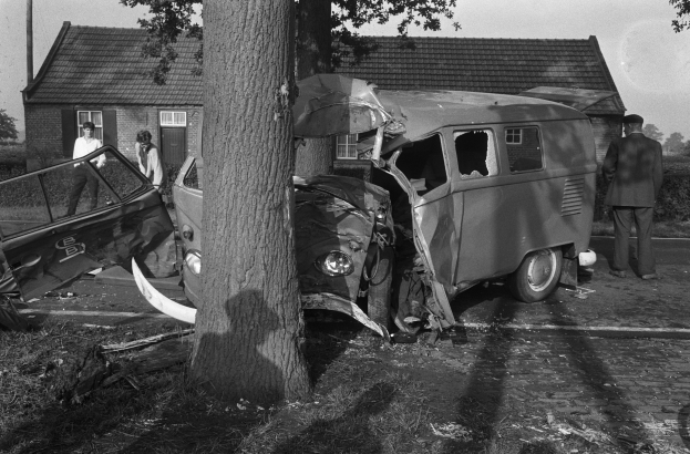 Schwarz-weißes Bild eines verunglückten Autos am Straßenrand mit einem Baum im Vordergrund, einige Menschen in der Nähe und Häuser, Bäume und ein Mast im Hintergrund.