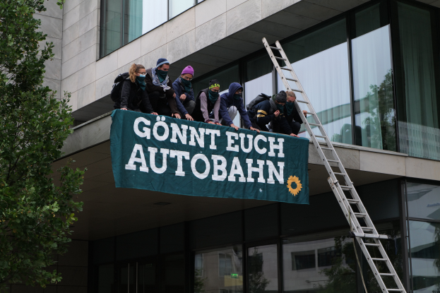 Eine Gruppe von Menschen steht auf dem Dach eines Gebäudes, hält eine Fahne hoch, mit einem Baum links und Glasfenstern im Hintergrund, die scheinbar eine Demonstration für die deutsche Regierung unterstützen.