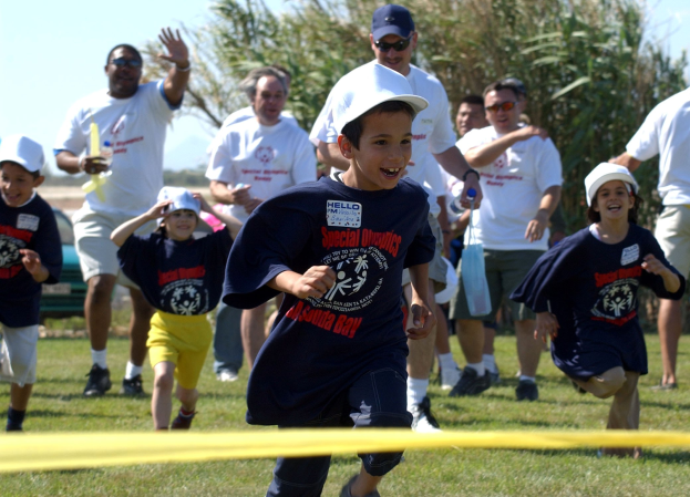 Kinder bei einem Lauf auf einer grünen Wiese mit einem gelben Band im Vordergrund, einige tragen Mützen und Schutzbrillen, andere halten Gegenstände, mit Bäumen und einem klaren blauen Himmel im Hintergrund.