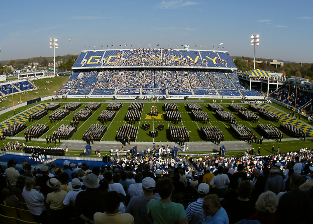 Eine große Menschenmenge in einem Stadion bei einem Fußballspiel, einige stehen, andere sitzen, auf einem Rasen unter einem klaren blauen Himmel, mit Laternen, Bäumen und Gebäuden im Hintergrund.