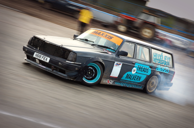 Ein Auto rast auf einer Rennstrecke, wobei Rauch ausstößt, mit anderen Fahrzeugen und Menschen im Hintergrund, alles durch die hohe Geschwindigkeit des Autos verwischt.